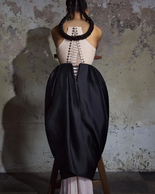 Back view of an adult female fashion model wearing a tan top and large black skirt. The model has braided hair and is in a studio.