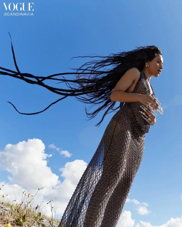 Woman in a sheer embellished dress with long dark braided hair flowing against a bright blue sky.