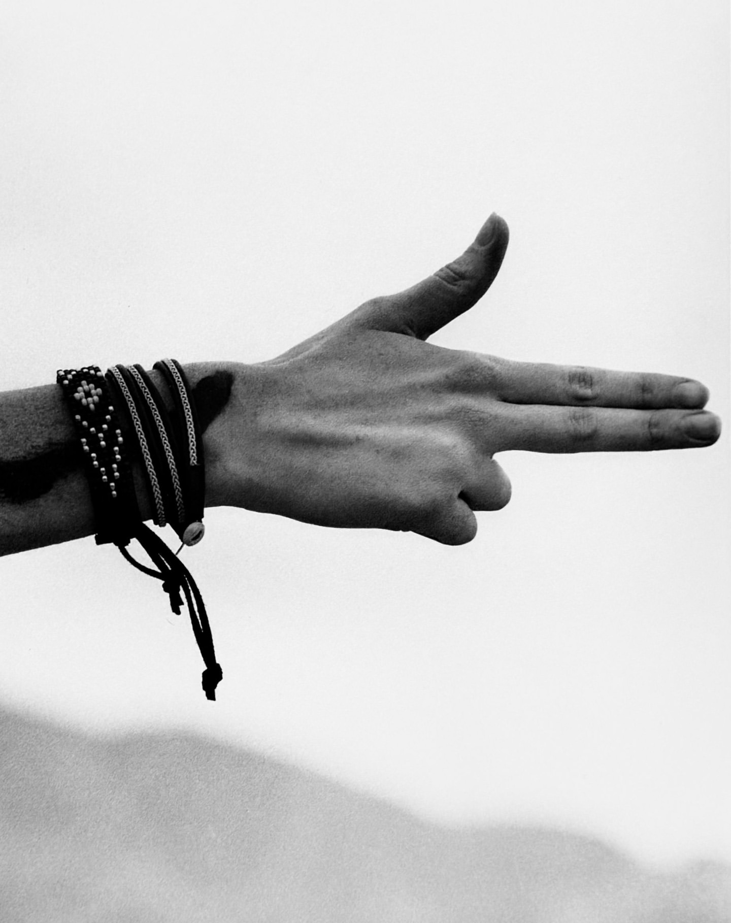 Black and white shot of an arm and hand making a gun gesture, accessorized with multiple stacked bracelets against a soft sky background.