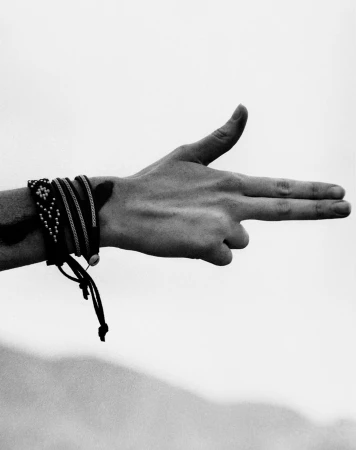 Black and white shot of an arm and hand making a gun gesture, accessorized with multiple stacked bracelets against a soft sky background.