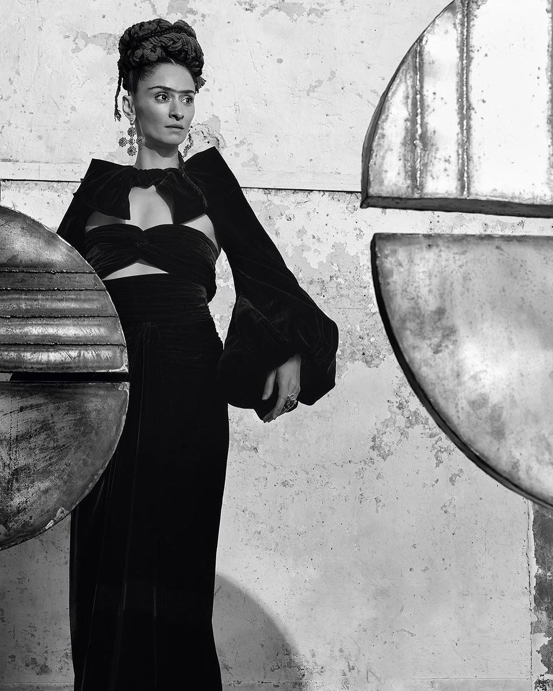Monochromatic portrait of a woman resembling Frida Kahlo, wearing a modern black dress with metal art pieces, posed against a distressed wall.