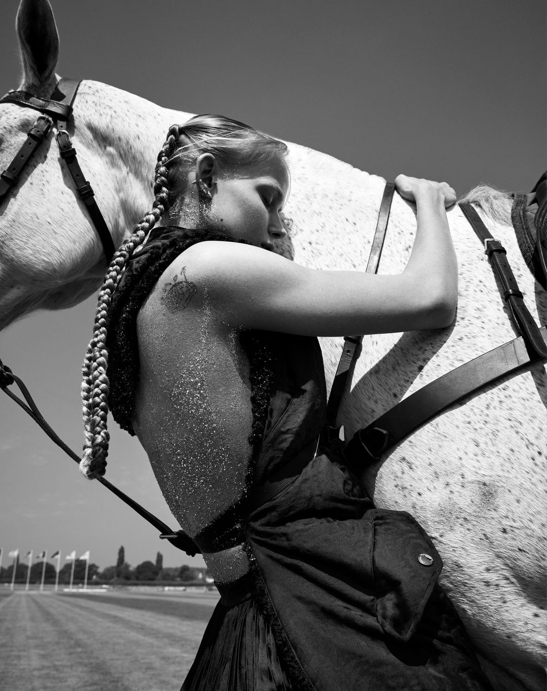 Black and white close-up of a woman with braided hair embracing a dappled grey horse with tack, against a blurred background.