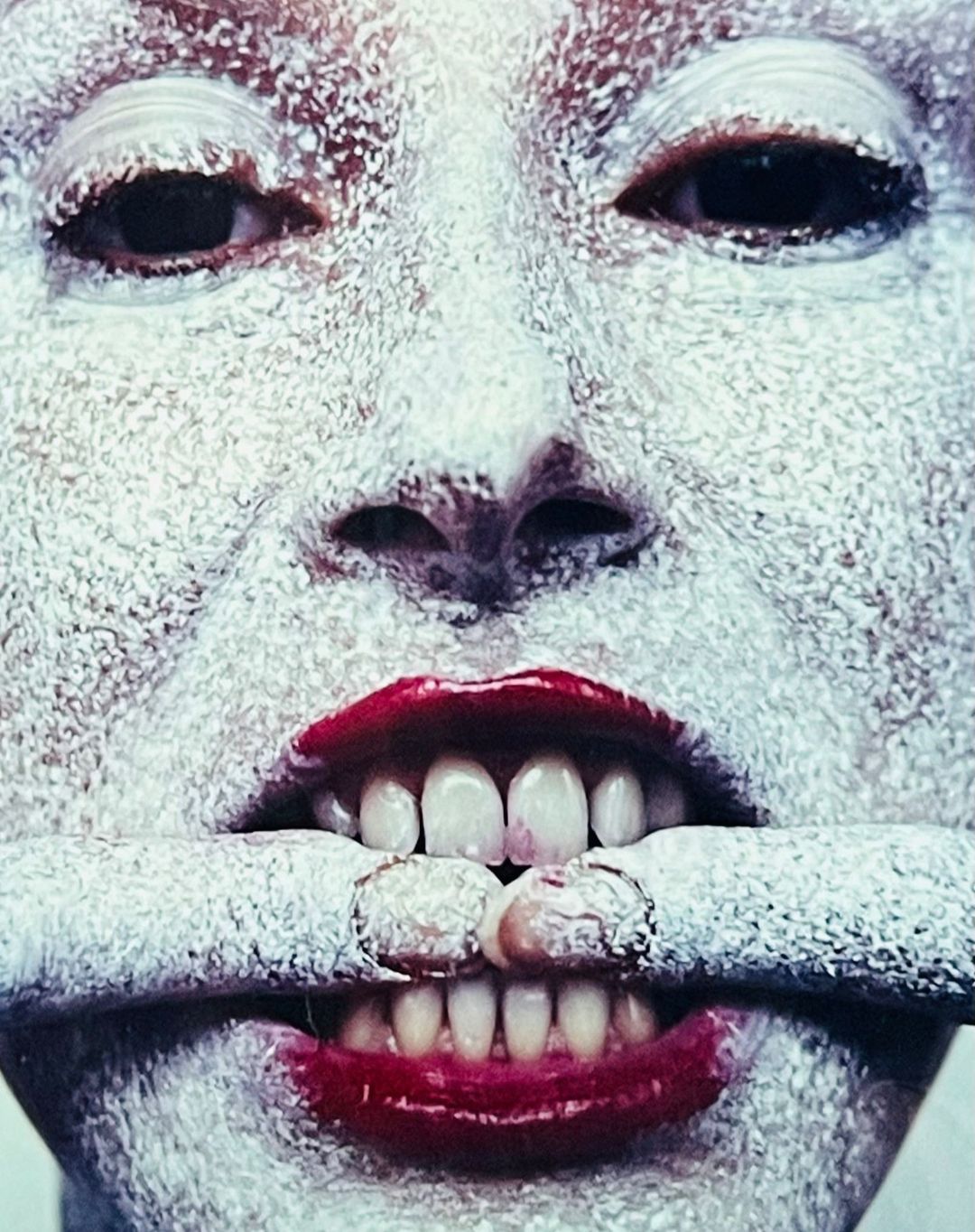 Close-up portrait of a person with white powder on their face and fingers holding lips apart to reveal teeth