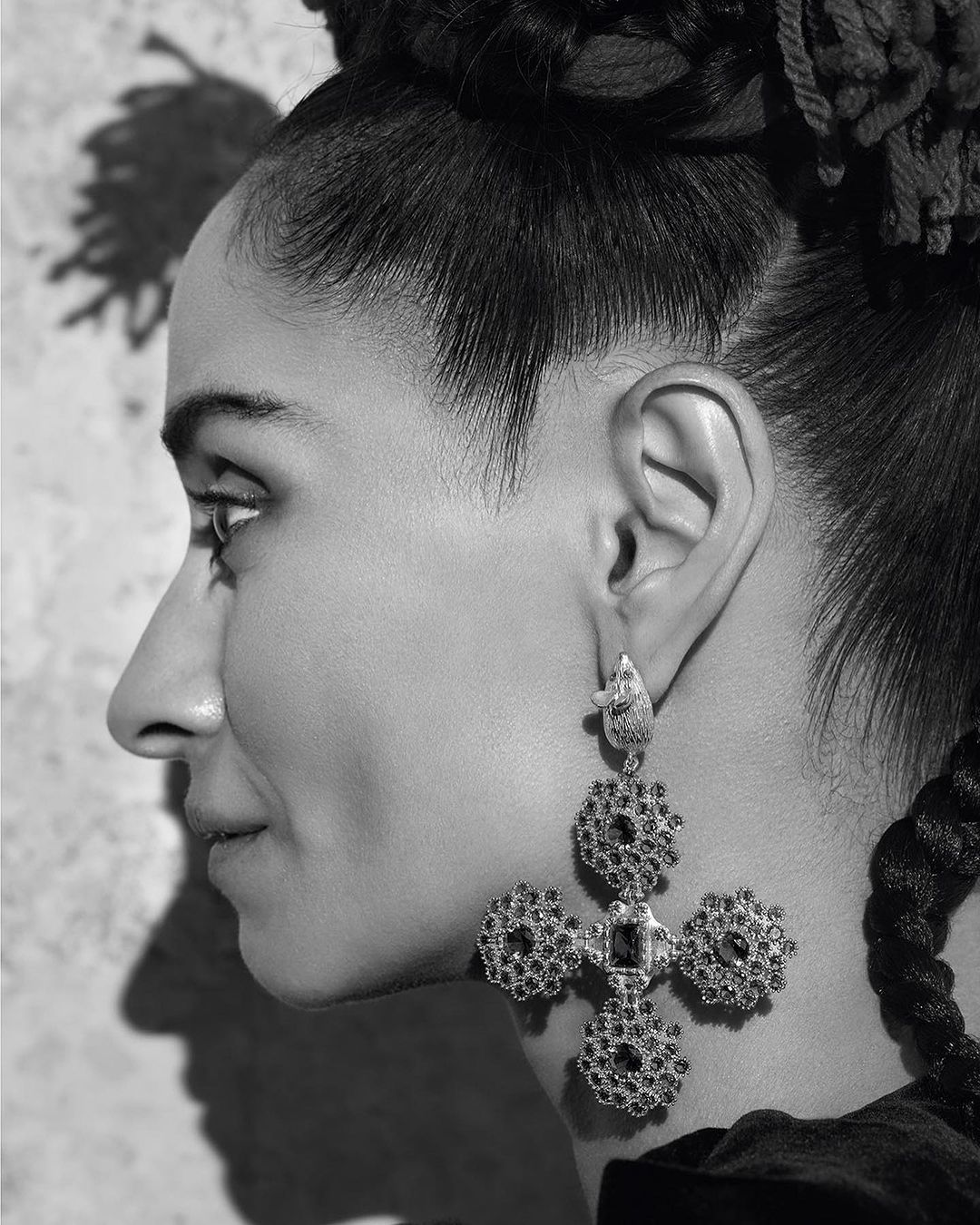 Profile of adult woman with braided hair wearing cross earrings in a studio with a textured backdrop in black and white.