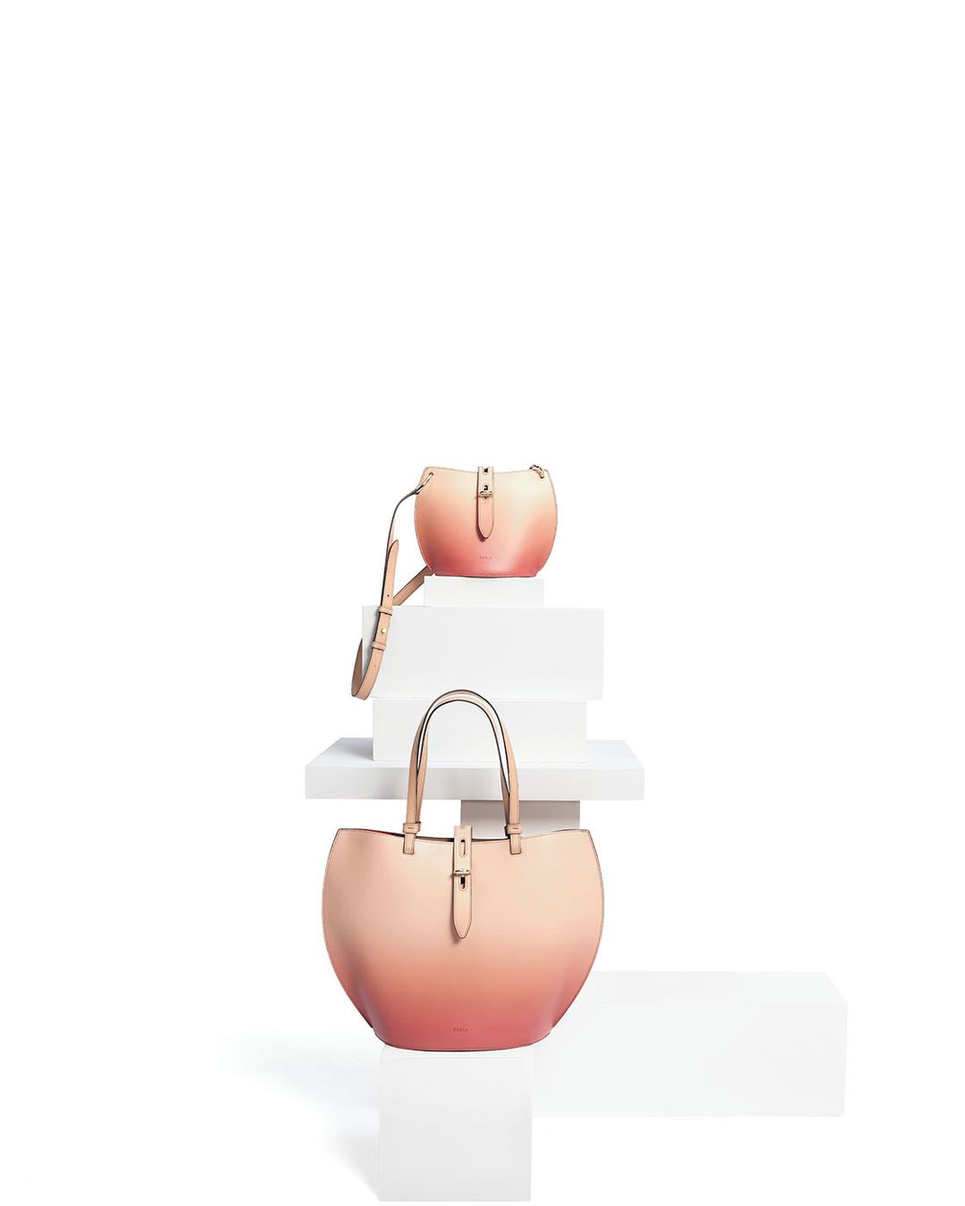 Two peach-ombre leather tote bags arranged on white steps in a product shot with soft studio lighting and a clean, minimalist composition.