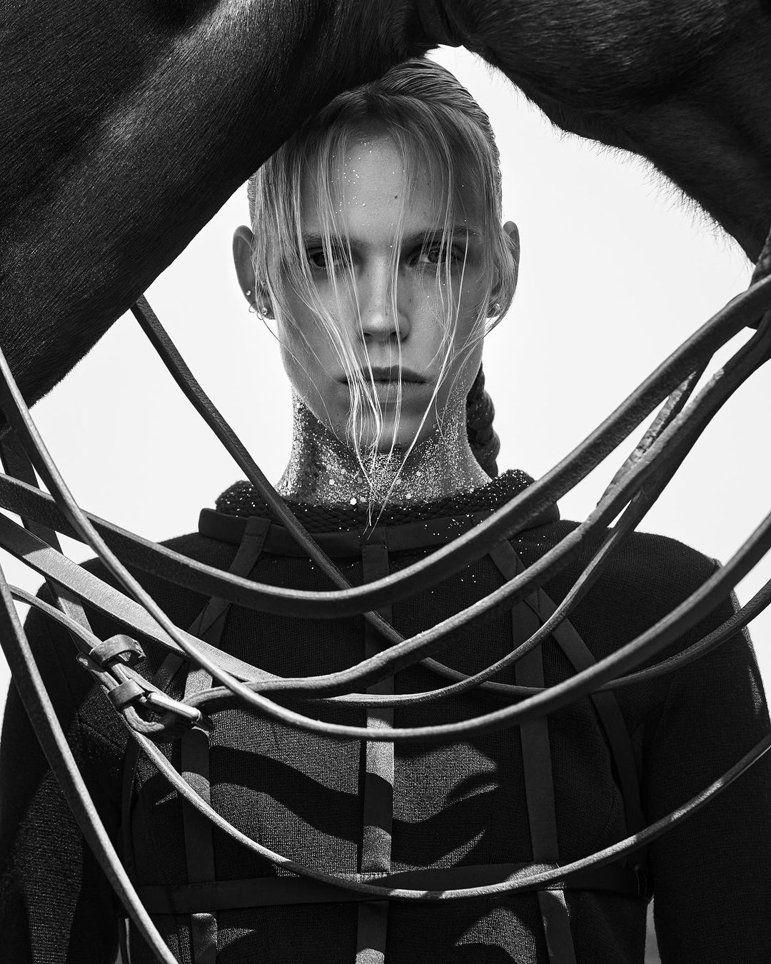 Black and white portrait of a young woman with braided hair and glittery collar, partially framed by a dark horse and reins.
