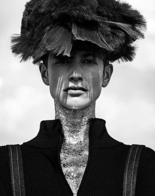 Monochrome portrait of a model with a distinctive dark feathered hat and sparkly glitter on the face and neck, set against a light sky.