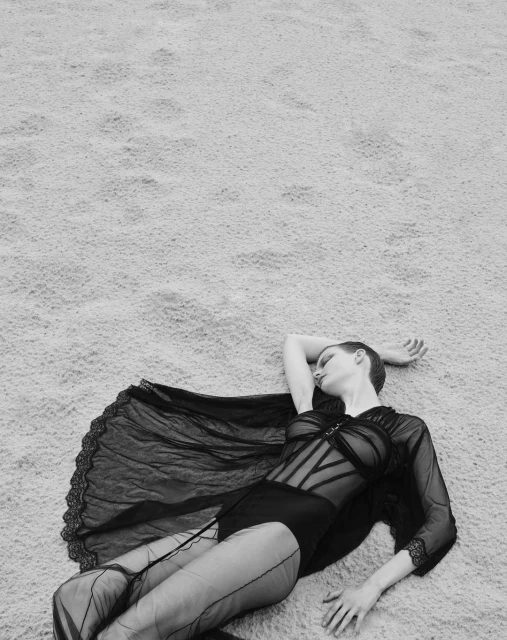 Black and white fashion portrait of a woman in sheer black clothing lying on a textured surface, showcasing editorial style and monochrome tones.