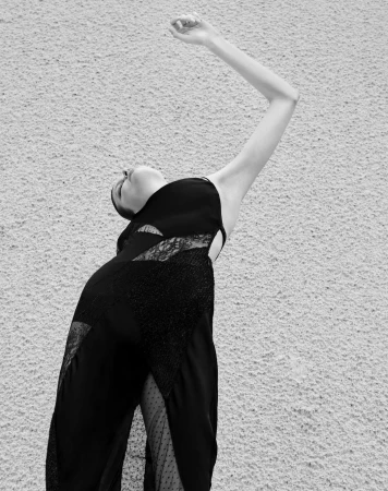 Black and white portrait of a woman in a dark dress arching backwards against a textured wall by Koto Bolofo
