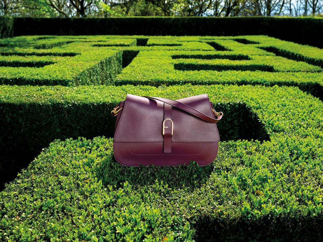 A dark red leather satchel handbag is centrally placed on a maze-like shrub, set in a lush green garden with trimmed hedges and dappled light.