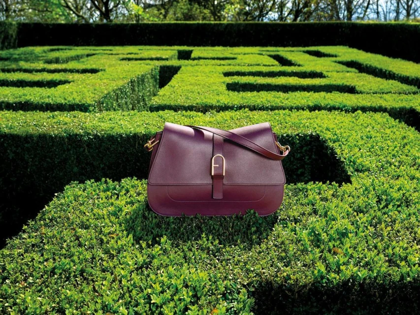A dark red leather satchel handbag is centrally placed on a maze-like shrub, set in a lush green garden with trimmed hedges and dappled light.