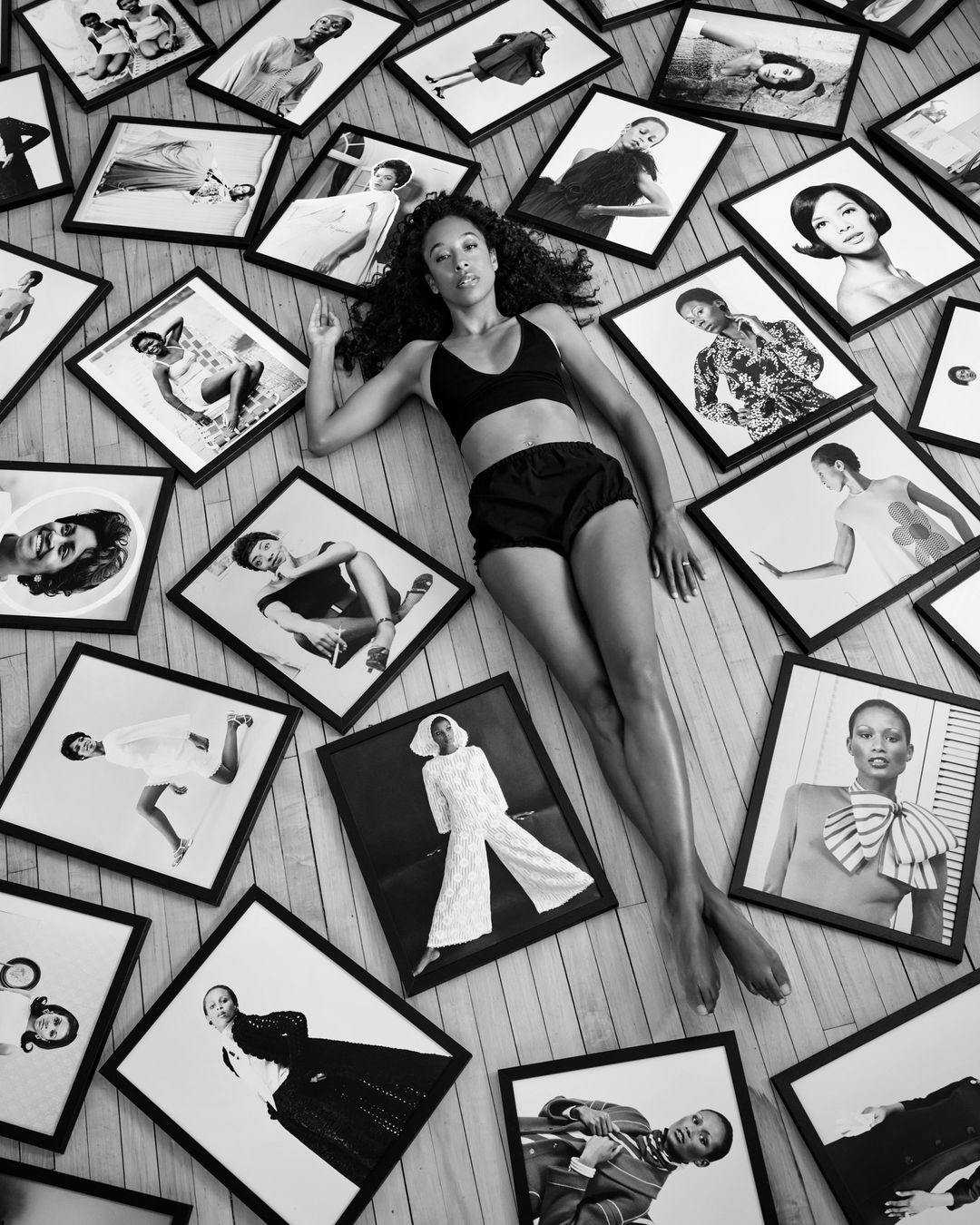 Black and white portrait of an African-American woman laying on a wooden floor surrounded by framed images of women.