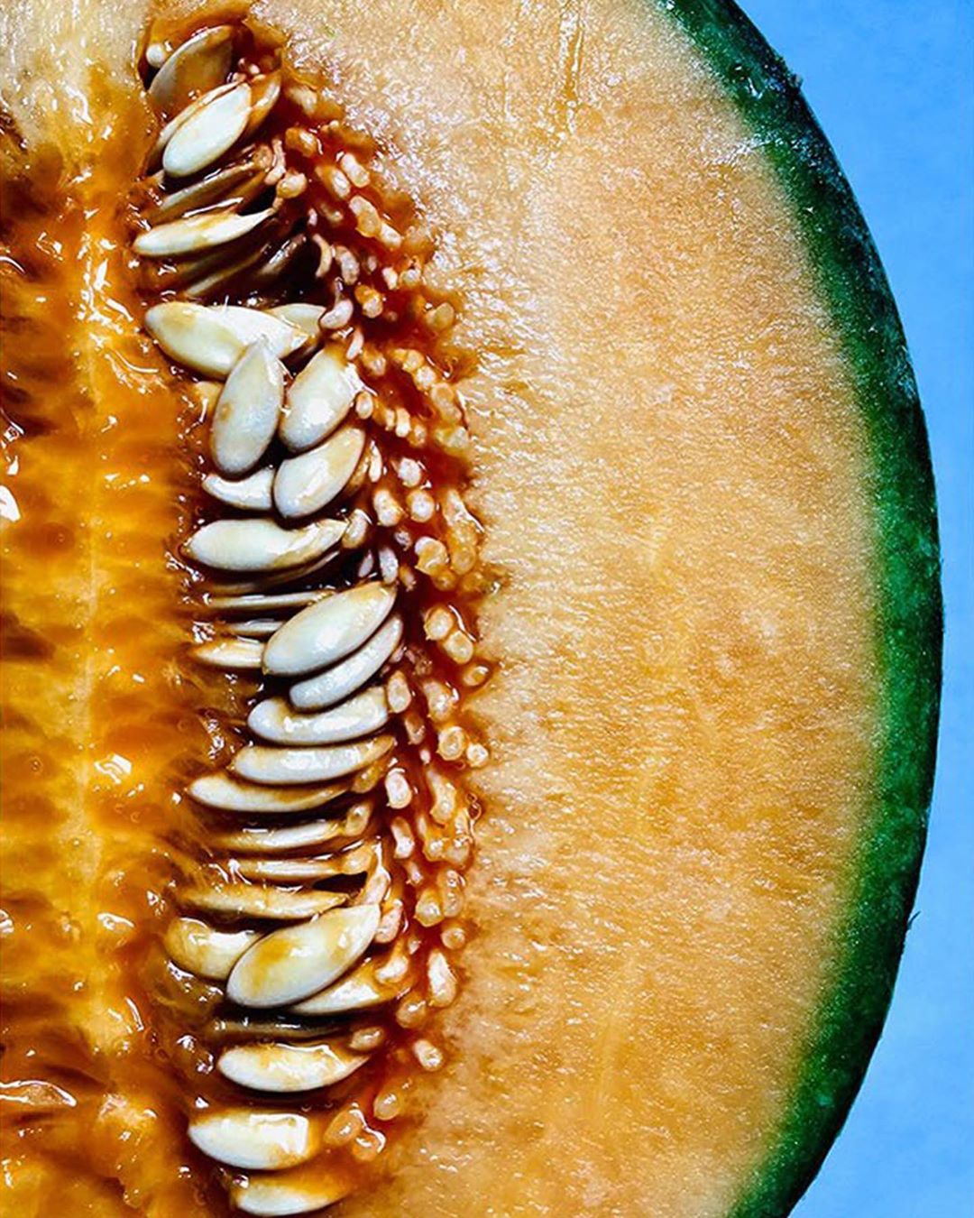 Close-up of a halved cantaloupe melon with seeds, vibrant orange flesh, and green rind, presented against a bright blue background.
