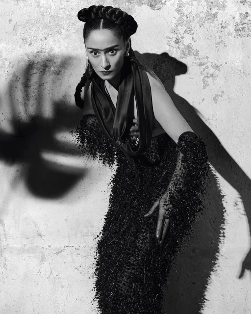 Striking black and white portrait of a woman with braided hair, wearing an ornate beaded gown, posing against a textured wall.