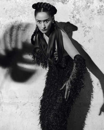 Striking black and white portrait of a woman with braided hair, wearing an ornate beaded gown, posing against a textured wall.