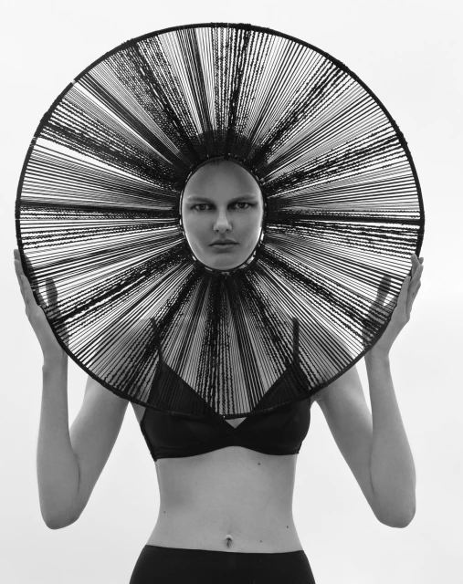 Monochromatic studio portrait of a bald female model wearing a geometric string headpiece and posing against a bright white backdrop.