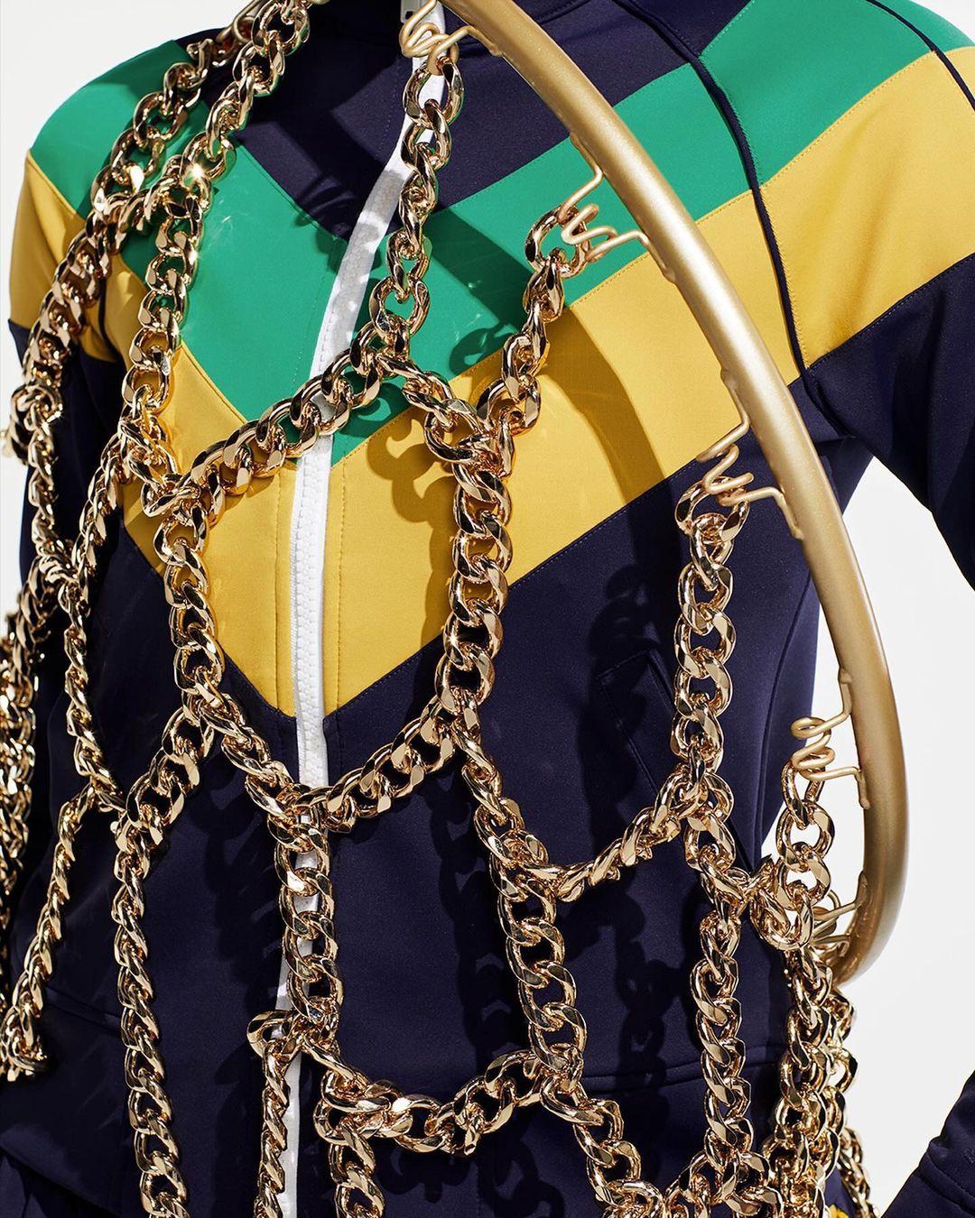 Cropped shot of a person in a color-blocked jacket, draped with chunky gold chains and a gold hula hoop, against a white backdrop.