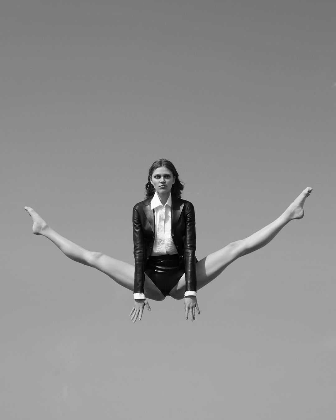 Black-and-white portrait of a female adult doing the splits in mid-air against a plain sky wearing a shirt, leotard, and blazer.