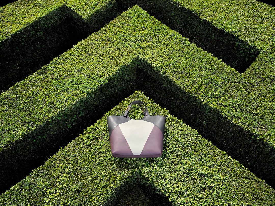 A luxury tote bag in shades of gray, white, and purple sits centered within a geometric boxwood maze on a sunny day, product shot.