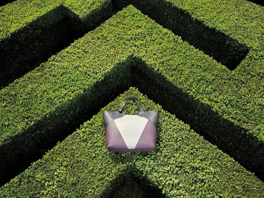 A luxury tote bag in shades of gray, white, and purple sits centered within a geometric boxwood maze on a sunny day, product shot.