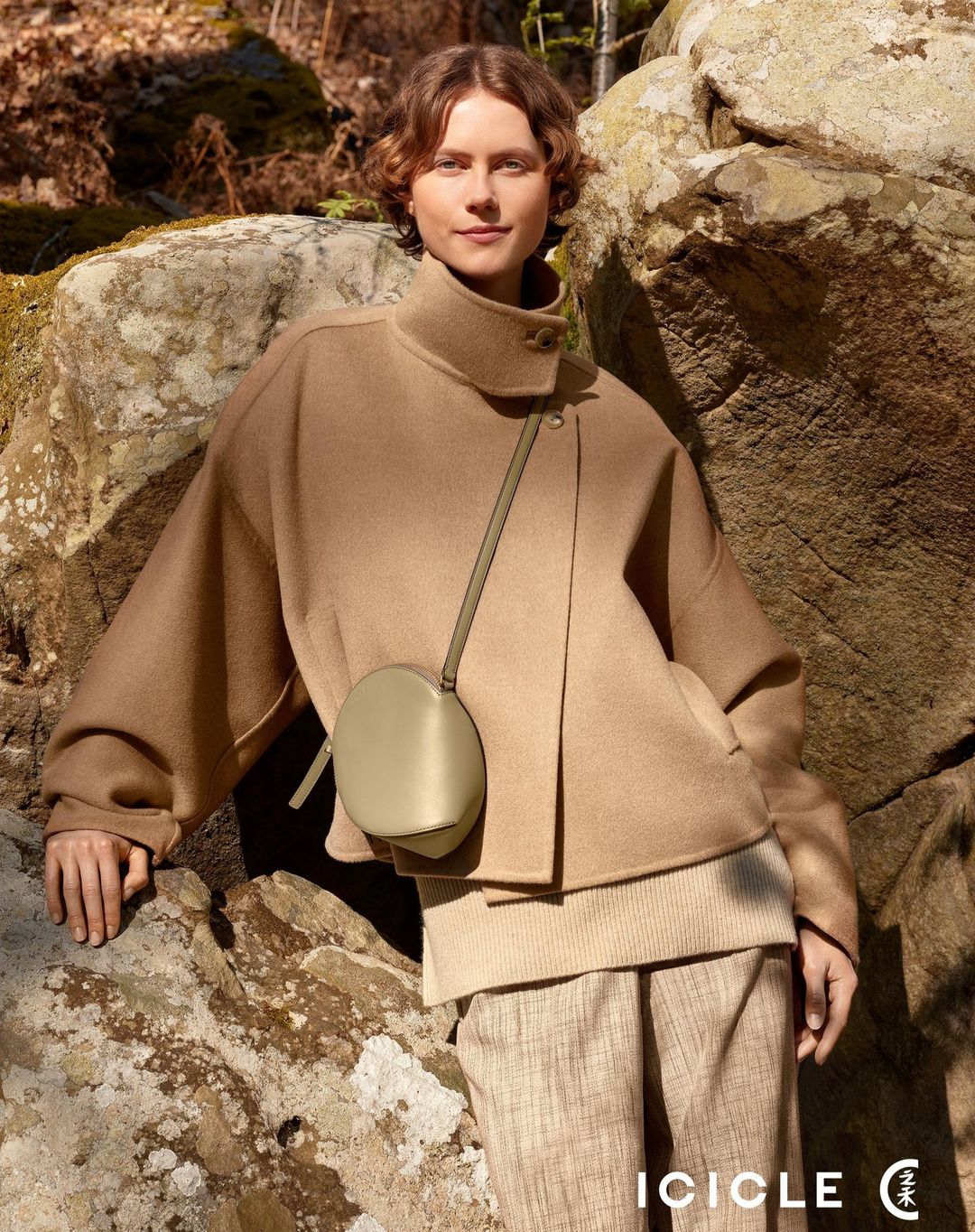 Woman in neutral-toned wool jacket and trousers poses near lichen-covered rocks outdoors, showcasing fashion and nature.