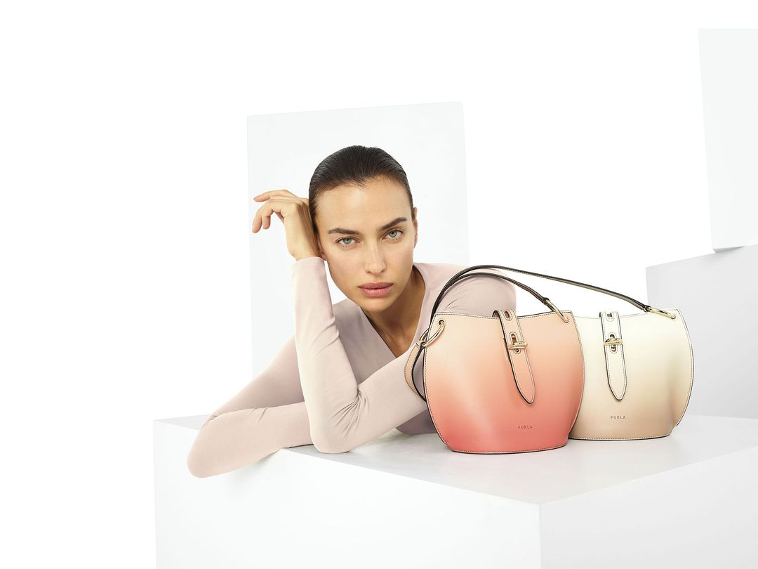 Fashion shot of a woman in front of two Furla handbags, one ombre peach to ivory, the other cream, against a bright white background.