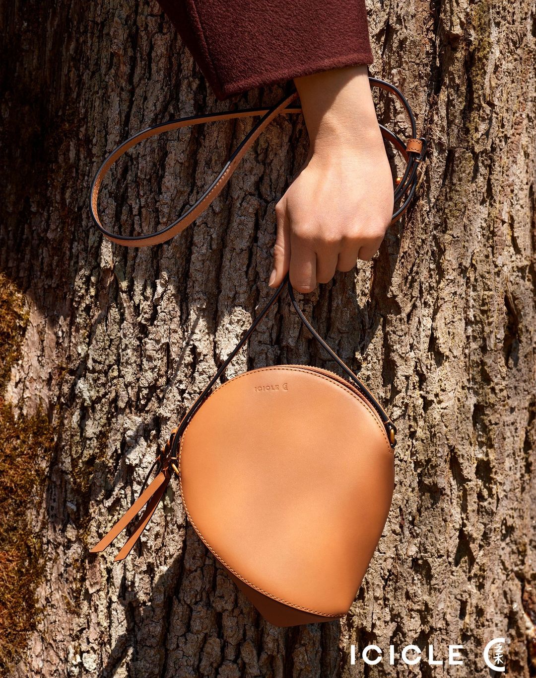 Tan leather crossbody bag by ICICLE is held by a woman in front of a textured tree trunk in a fashion product shot.