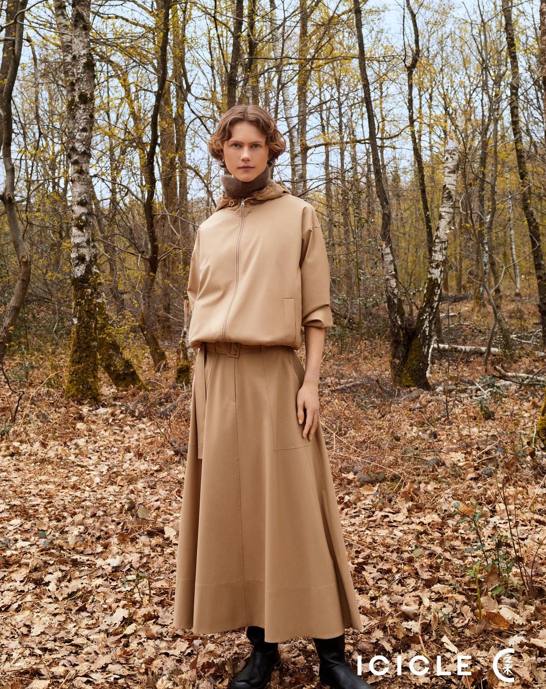 A woman in a tan outfit poses in a forest setting with birch trees and fallen leaves on the ground, displaying a minimalist fashion aesthetic.