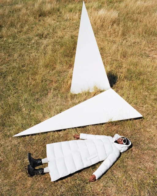 Woman in white puffer coat lying in a grassy field with large white triangular shapes in an outdoor fashion portrait.