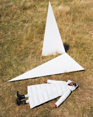 Woman in white puffer coat lying in a grassy field with large white triangular shapes in an outdoor fashion portrait.