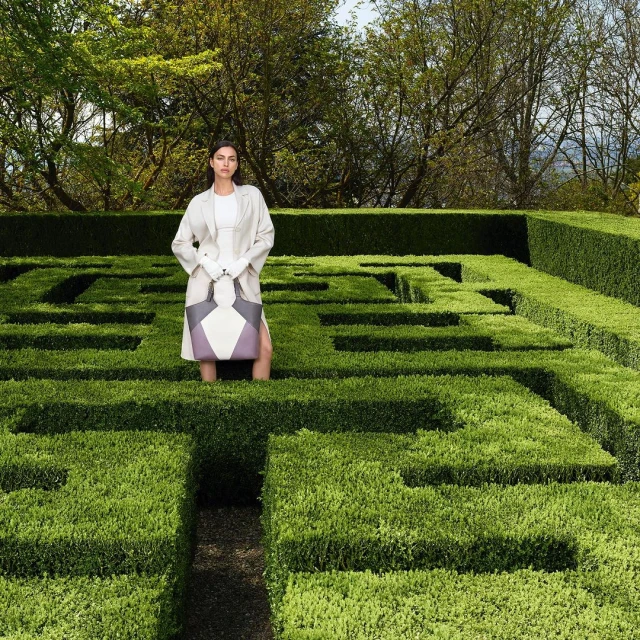 Adult female fashion model poses with a geometric tote bag in a green boxwood maze on an overcast day in a natural outdoor setting.