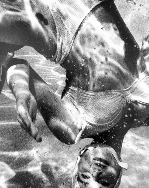 Monochrome underwater shot features a woman in a swimsuit and swim cap, her face and body distorted by the water's surface.