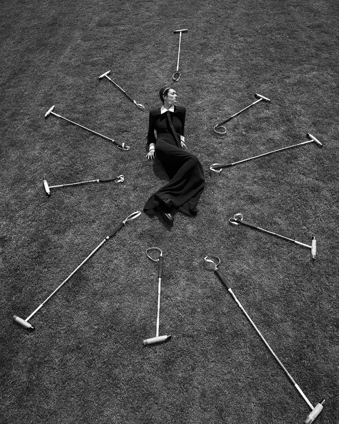 High-angle shot of a woman in a black dress reclining on grass surrounded by polo mallets in a radial pattern, monochrome.