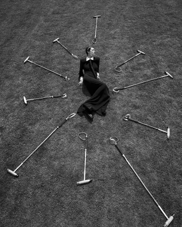 High-angle shot of a woman in a black dress reclining on grass surrounded by polo mallets in a radial pattern, monochrome.