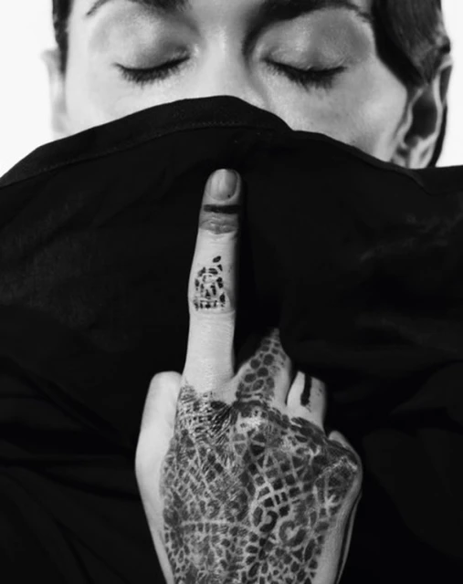 Monochromatic portrait of a person with eyes closed, part of their face covered by a fabric with patterned henna tattoo on hand.