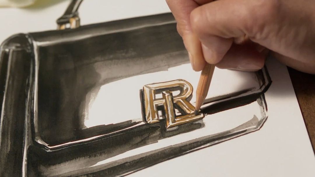 Close-up of a fashion handbag drawing in progress, with a hand using a peach colored pencil to add detail to the design's metal logo.