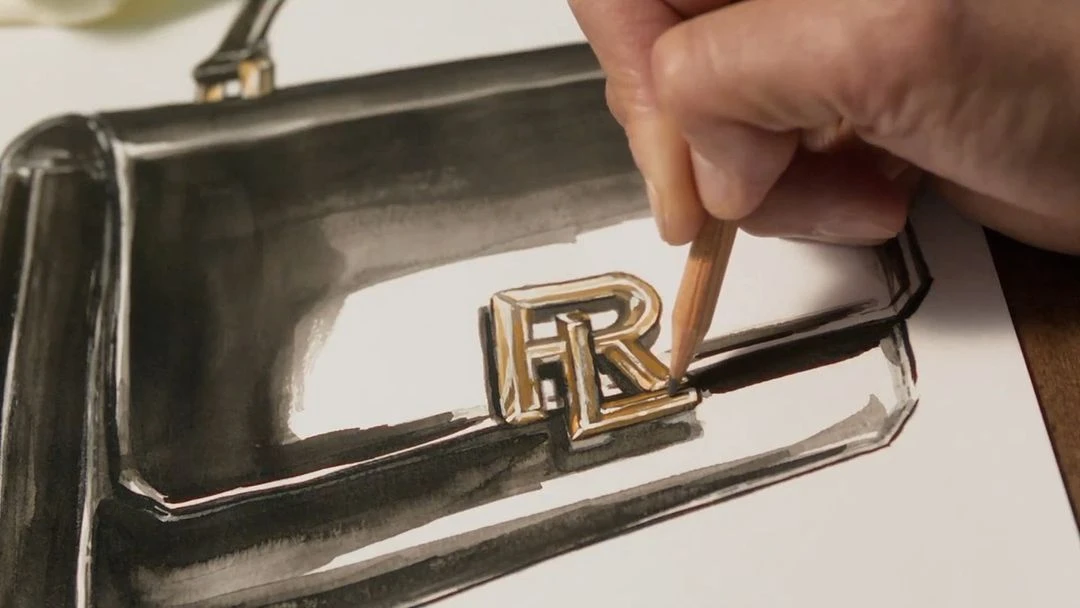 Close-up of a fashion handbag drawing in progress, with a hand using a peach colored pencil to add detail to the design's metal logo.