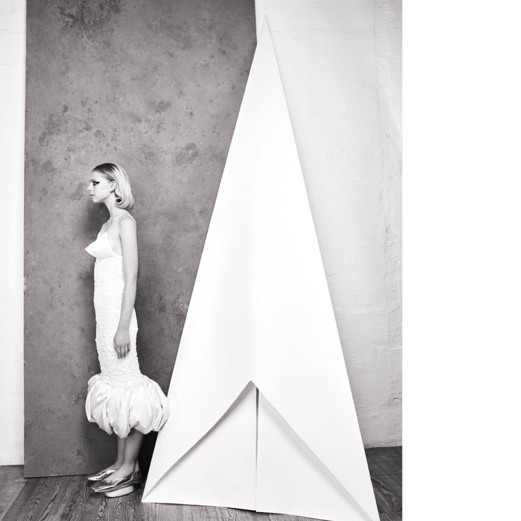 Woman in a couture white dress stands in profile next to a paper sculpture in a minimalist studio with gray and white walls.