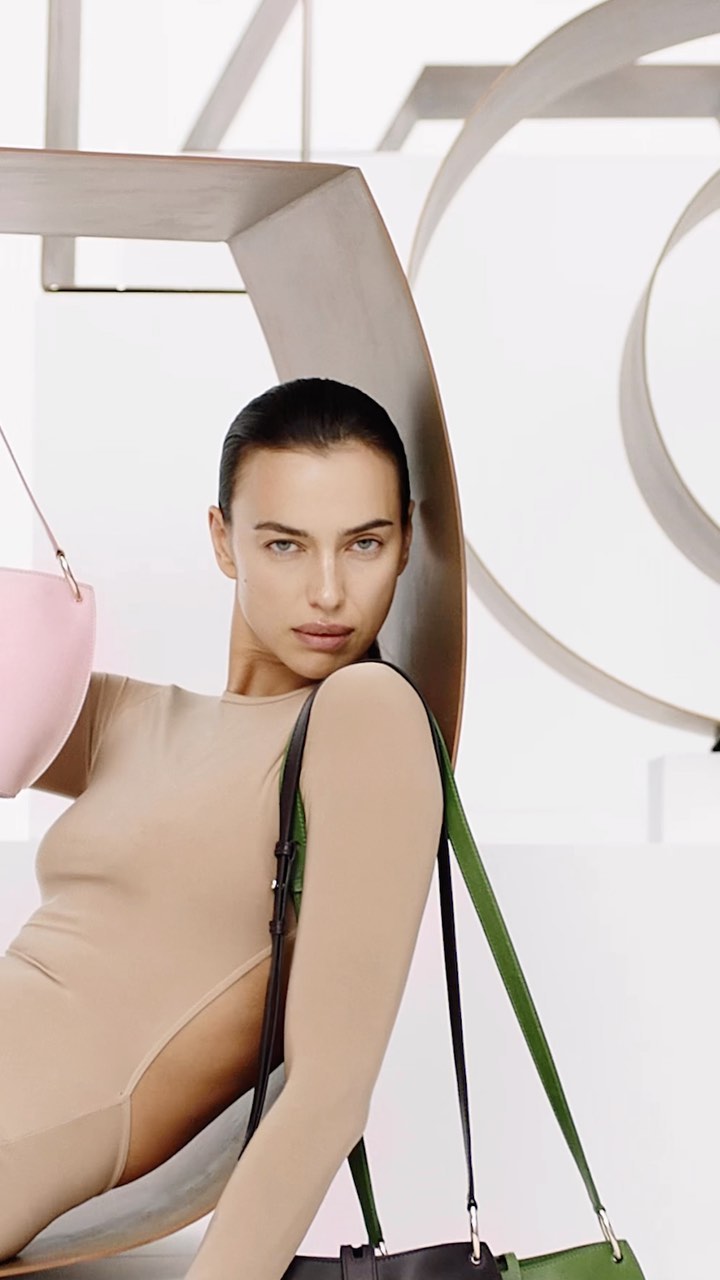 Indoor shot of a female model in a neutral bodysuit, holding a pink handbag and a green and black tote bag, posing in a photo studio.