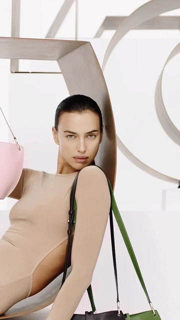 Indoor shot of a female model in a neutral bodysuit, holding a pink handbag and a green and black tote bag, posing in a photo studio.
