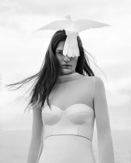 Black and white portrait of a woman with a white dress, sheer top, and a white dove perched on her head against a bright background.