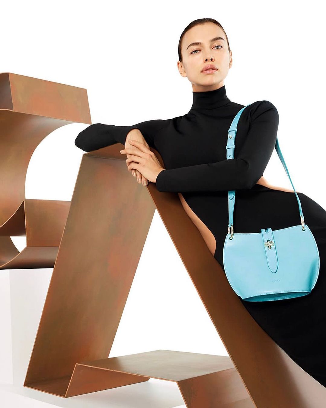 Adult woman in a black turtleneck dress poses with a light blue leather shoulder bag on a geometric bronze-colored metal sculpture indoors.