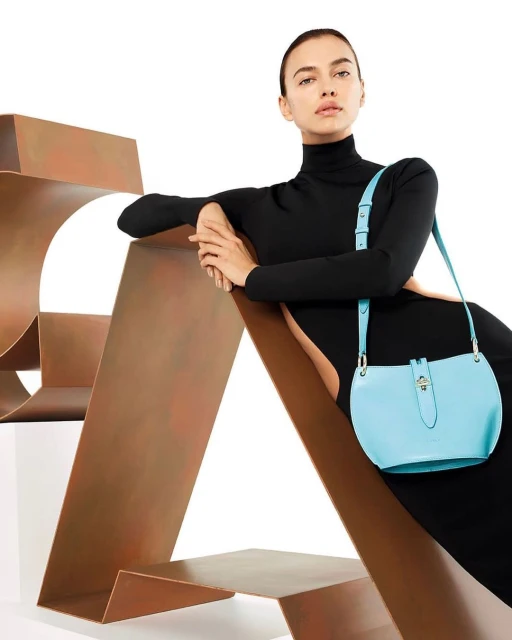 Adult woman in a black turtleneck dress poses with a light blue leather shoulder bag on a geometric bronze-colored metal sculpture indoors.