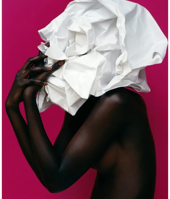 Striking studio portrait of a Black woman with dark skin, holding her hands up towards a large white paper sculpture on her head.