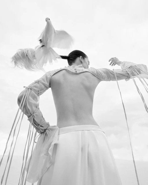 Monochrome image of a person in flowing white garments with a white dove perched on their head, viewed from the back in an outdoor setting.