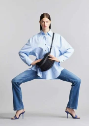 Model in wide stance wearing oversized light blue button-down shirt and blue jeans with a black crossbody bag