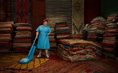An older woman in a blue dress vacuums a large patterned rug inside a room filled with towering stacks of oriental carpets.