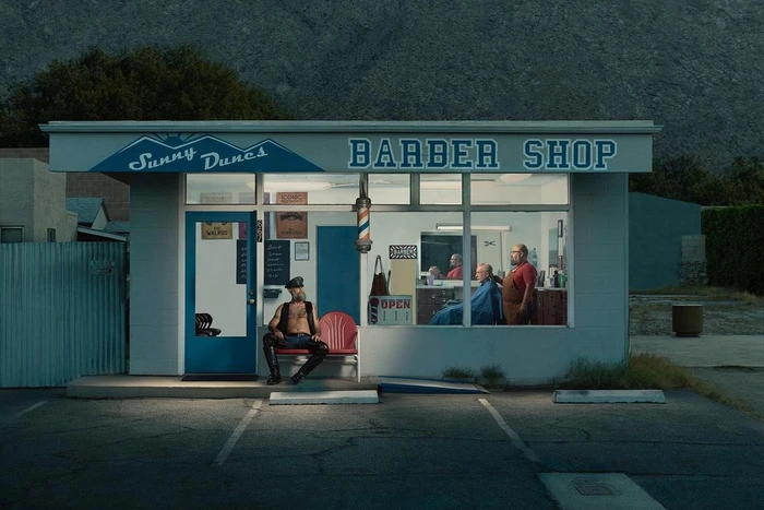 A man in black leather sits outside a barbershop named Sunny Dunes while another man gets a haircut inside.