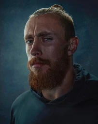 Portrait of a man with a reddish-brown beard and hair in a top knot, wearing a dark hoodie against a textured blue background.