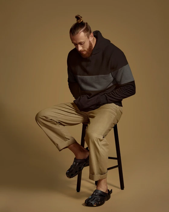Man with top knot and beard wearing a dark hoodie and khaki trousers seated on a stool against a warm tan background.
